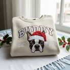 Felpa personalizzata color crema con ritratto del cane ricamato a colori e cappello di Natale, nome “BUDDY” e dettaglio “EST. 2025”.
