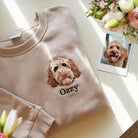Felpa personalizzata con ricamo del ritratto di un cane in stile full color e il nome “Ozzy”, realizzata a partire da una foto del pet. Felpa beige piegata su un tavolo con la foto originale dell’animale accanto.

