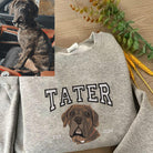 Felpa grigia personalizzata con ricamo a colori del ritratto di un cane, realizzato a partire dalla foto del cane mostrata nell’angolo superiore sinistro; design con il nome “Tater” e l’anno “2024”.
